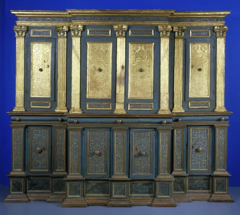Large ornate cabinet with blue-green and gold painted panels, featuring tall vertical doors with gilded designs, decorative columns, and multiple drawers along the bottom, set against a blue background.