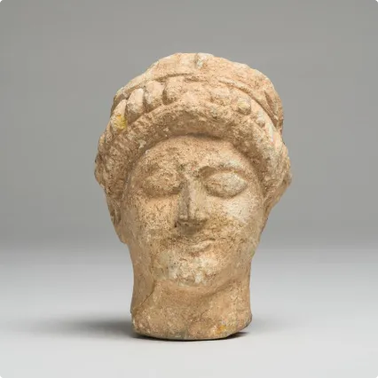 stone sculpture of a woman’s head with stylized features, closed eyes, and braided hair, displayed against a neutral gray background.