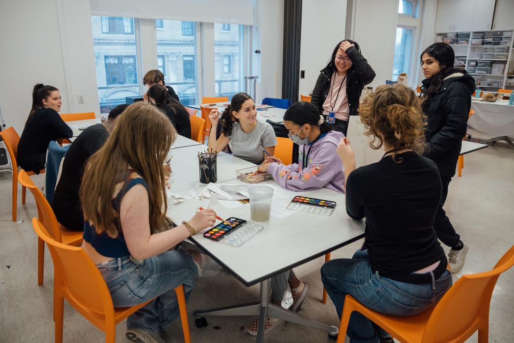 Photograph of teen girls and boys seated at a white table with orange chairs, doing a watercolor art project.