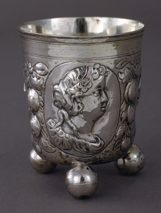 Silver metal cup with straight sides and three rounded feet. On the side, a woman’s face with curly hair and other decorative elements are rendered in 3D form.