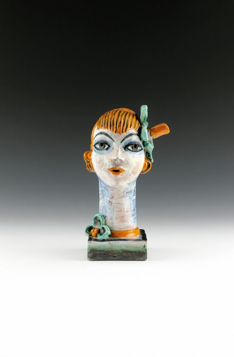 Ceramic bust of a doll-like woman with light skin, blond hair, red lips, and rouged cheeks. Her eyes and eyebrows are outlined thickly with blue pigment and she wears a hat tipped to the right with a green flower tucked under the brim.